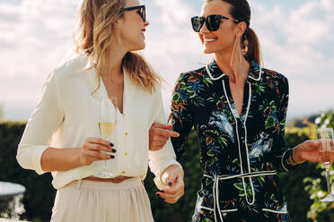 Fashionable female friends with wine walking outdoors looking at each other and smiling. Two stylish women walking together with glass of wine. - JLPSF24035