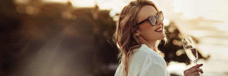 Glamorous woman having wine outdoors with large copyspace background. Beautiful female model wearing sunnglasses with a glass of wine. - JLPSF24050