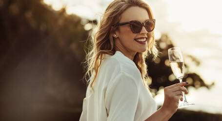 Elegant woman in sunglasses with a glass of wine outdoors. Smiling caucasian female having wine and looking backwards. - JLPSF24051