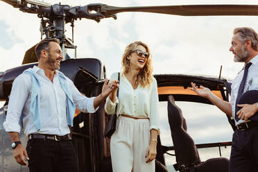 Smiling man and woman alighted from a private helicopter with pilot standing by. Couple getting off a private aircraft with mature pilot. - JLPSF24062