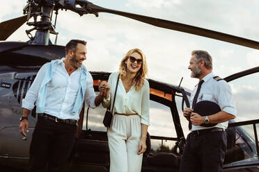 Beautiful woman escorted by her boyfriend out off a private helicopter with pilot standing by. Smiling couple traveling by their private helicopter. - JLPSF24063