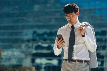 Asian businessman walking outdoors using his smart phone. Man reading text message on his mobile phone while walking outdoor against building. - JLPSF24083