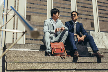 Two business partners sitting on the steps outdoors and talking. Korean business men discussing work while sitting on stairs outside in the city. - JLPSF24092