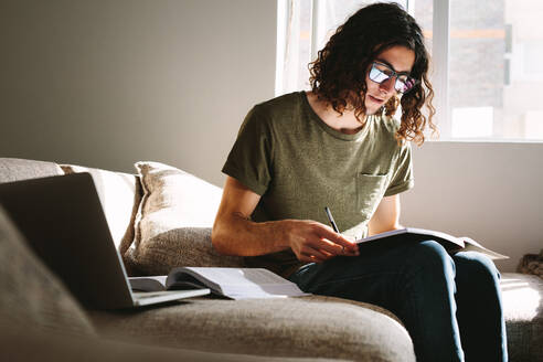 Student studying at home with books and laptop sitting on couch. Man preparing for college exams. - JLPSF24095
