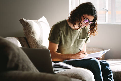 Young man studying for exams sitting on couch at home. Student studying at home with books and laptop. - JLPSF24096