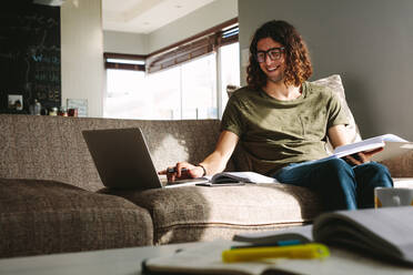 Smiling young man studying for exams sitting on couch at home. Student studying at home with books and laptop. - JLPSF24098