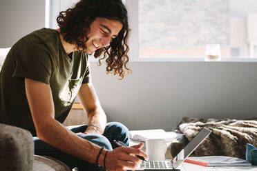 Side view of a student studying at home with a laptop on table. Cheerful student sitting at home beside a window and studying. - JLPSF24103