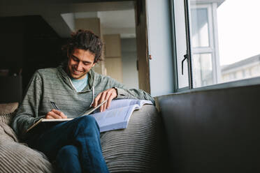 Smiling man sitting comfortably at home writing in a book. Student writing notes from a text book sitting on couch beside a window. - JLPSF24109
