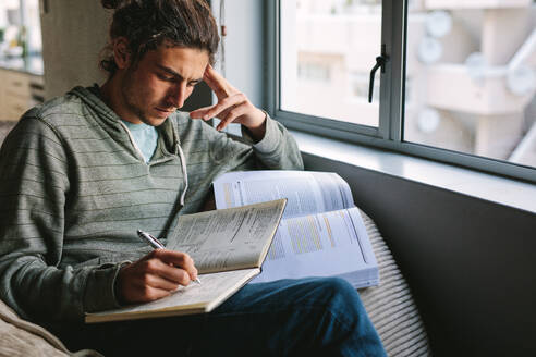 Student doing his home work sitting on couch beside a window. Man writing in book sitting at home with focus and concentration. - JLPSF24110