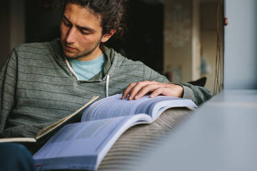 Student making notes from a reference book sitting on a couch. Young man studying seriously sitting at home. - JLPSF24111