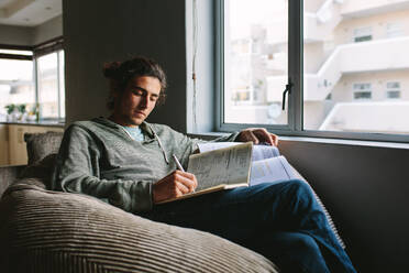 Young man writing notes sitting comfortably on a couch beside a window. Student studying and preparing notes sitting at home. - JLPSF24112