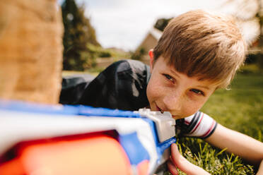 Kid in cape hiding behind a wooden log with toy gun in hands. Cute boy playing with toy gun in playground. - JLPSF24127