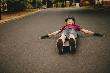 Small boy lying on his skateboard using his hands to guide himself along road. Playful boy skateboarding outdoors. - JLPSF24129