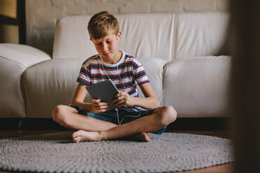 Young boy sitting on floor wearing earphones and playing video games on digital tablet. Boy in headphones playing online game on tablet at home. - JLPSF24142