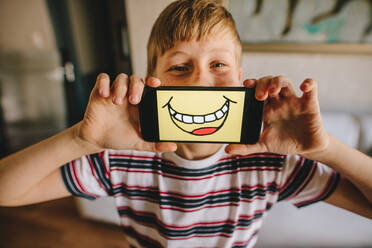 Boy holding a smartphone in front of his face with smiley picture on the display. Boy pretending to be happy at home. - JLPSF24147