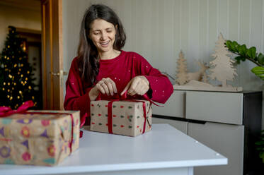 Glückliche Frau bindet Schleife an Weihnachtsgeschenke zu Hause - ANAF00323