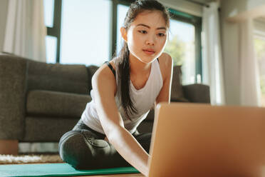 Frau in Sportkleidung, die auf dem Boden sitzt und einen Laptop benutzt. Fitness-Frau, die zu Hause auf dem Laptop Lektionen über spirituelle Praktiken betrachtet. - JLPSF24149