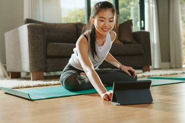 Lächelnde Frau sitzt auf einer Trainingsmatte und schaut sich Trainingsvideos auf einem digitalen Tablet an. Junge chinesische Frau in Sportkleidung trainiert und schaut sich Online-Videos auf einem Tablet-PC an. - JLPSF24156