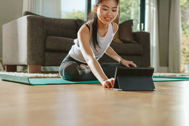 Woman using digital tablet while exercising at home. Female in sportswear sitting on floor and watching exercise video for her workout. - JLPSF24157