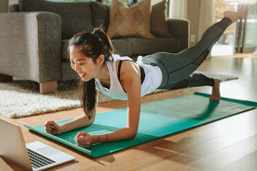 Smiling woman exercising at home and watching training videos on laptop. Chinese female doing planks with a leg outstretched and looking at laptop. - JLPSF24159