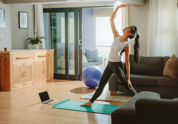 Frau, die in ihrem Wohnzimmer trainiert, steht auf einer Yogamatte und dehnt sich mit einem Laptop auf dem Boden. - JLPSF24163