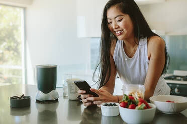 Woman leaning to kitchen counter using mobile phone in morning. Beautiful young woman standing by kitchen counter with bowl of fresh fruits and reading text message on her cell phone. - JLPSF24169