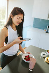 Frau, die ihre gesunde Frühstücksschüssel in der Küche fotografiert. Chinesische Frau, die ein gesundes Frühstück mit einem Mobiltelefon fotografiert - JLPSF24172