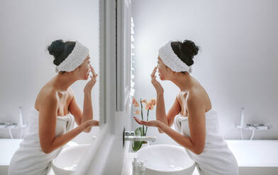Frau, eingewickelt in ein Handtuch, cremt sich vor einem Spiegel ein. Asiatische Frau bei der Körperpflege im Badezimmer. - JLPSF24175