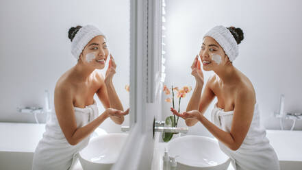 Female in towel applying cream onto her face. Asian woman in bathroom looking in to the mirror and applying cream on her face. - JLPSF24176