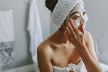 Junge asiatische Frau im Badezimmer, die eine Gesichtsmaske auf ihr Gesicht aufträgt. Eine Frau, die eine Gesichtsmaske auf ihr Gesicht aufträgt. - JLPSF24178