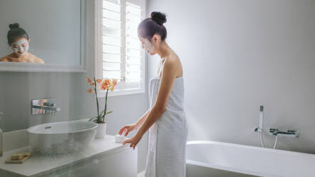 Asiatische Frau in Handtuch eingewickelt steht im Badezimmer und trägt Feuchtigkeitscreme auf. Frau mit Gesichtsmaske trägt Körperlotion auf. - JLPSF24183