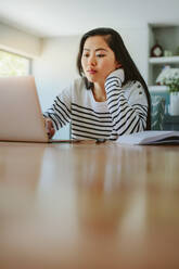 Frau sitzt zu Hause am Tisch und arbeitet am Laptop. Asiatisches Mädchen studiert zu Hause. - JLPSF24187