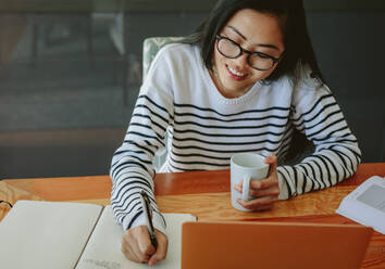 Frau schreibt Notizen in Tagebuch und hält eine Kaffeetasse in der Hand. Asiatische Frau studiert zu Hause mit Laptop auf dem Tisch. - JLPSF24189