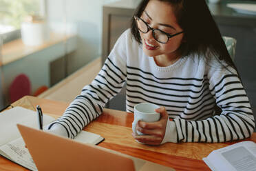 Eine Frau macht sich Notizen in ihrer Molkerei, hält eine Tasse Kaffee und einen Laptop vor sich auf dem Tisch. Eine asiatische Studentin arbeitet zu Hause an ihrer Aufgabe. - JLPSF24190