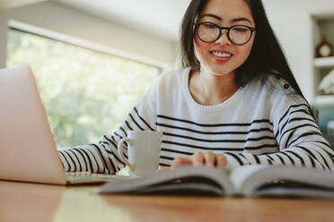 Lächelnde junge Frau liest ein Buch mit Laptop und Kaffeetasse auf dem Tisch. Asiatisches Mädchen mit Brille studiert zu Hause. - JLPSF24191
