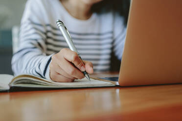 Nahaufnahme einer weiblichen Hand, die sich Notizen in einem Buch macht, mit einem Laptop auf dem Tisch: Studentin beim Lernen zu Hause. - JLPSF24192