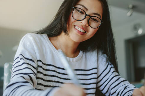 Lächelnde junge Frau mit Brille studiert zu Hause. Schöne asiatische Mädchen Studentin Schreiben und Lächeln drinnen. - JLPSF24193