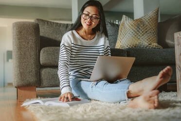 Frau sitzt auf dem Boden und liest ein Buch mit Laptop. Glückliche asiatische Studentin studiert zu Hause. - JLPSF24198
