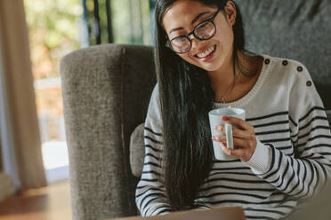 Lächelnde junge Frau mit Kaffee in der Hand und Laptop. Asiatische Frau entspannt sich zu Hause mit Laptop. - JLPSF24202