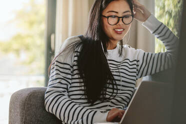 Frau mit Laptop-Computer auf dem Sofa sitzend und mit Kopfhörern. Lächelnde Frau, die sich zu Hause entspannt und am Laptop-Computer arbeitet. - JLPSF24203