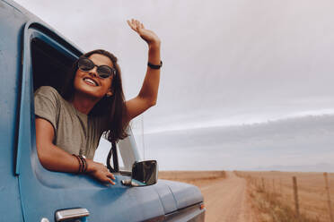 Happy woman enjoying road trip. Female traveling by a car looking out from window with her arm raised. - JLPSF24232