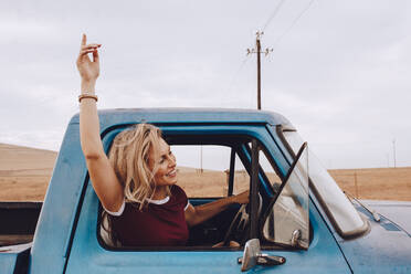 Beautiful young woman driving a car and enjoying road trip. Excited young female raising her hand out of the car while driving. - JLPSF24248