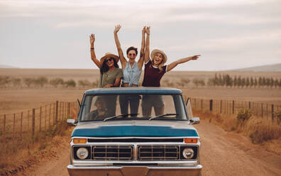 Eine Gruppe von Freundinnen, die in einem Pickup fahren und sich an einem Sommertag amüsieren. Drei Frauen stehen mit erhobenen Armen und lächelnd am Heck des Pickups. - JLPSF24259