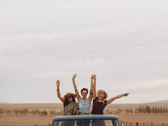 Eine Gruppe von Freunden auf einem Roadtrip durch die Landschaft. Drei junge Frauen stehen mit erhobenen Händen lachend auf dem Rücksitz eines offenen Lastwagens. - JLPSF24260