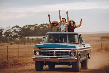 Drei Frauen auf einem Roadtrip durch die Landschaft reisen auf der Ladefläche eines Pickups. Freundinnen stehen mit erhobenen Händen lachend auf der Ladefläche eines offenen Trucks. - JLPSF24261