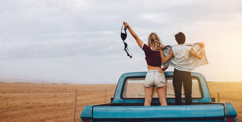 Rückansicht von zwei Frauen, die auf dem Rücksitz eines offenen Lastwagens stehen, während sie aufs Land fahren. Die Frau hält ihren BH und ihre Freunde stehen mit aufgeknöpftem Hemd da. - JLPSF24262