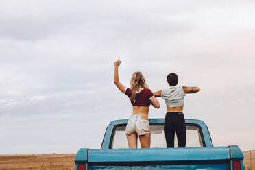 Rückansicht von zwei Frauen, die auf der Ladefläche eines Pickups stehen und ihre Hemden hochheben, während sie einen Ausflug aufs Land machen. - JLPSF24263