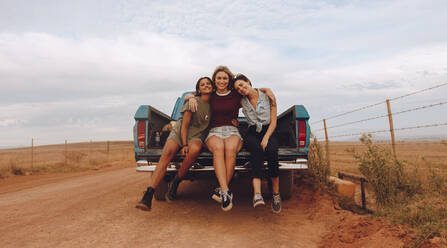 Porträt von drei jungen Freundinnen, die auf der Ladefläche eines Pickups sitzen, der auf einer Landstraße geparkt ist. Gruppe von Frauen auf einer Landpartie. - JLPSF24269