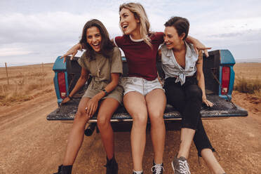 Three women traveling in a open back pickup truck through country road. Female friends having fun on their road trip. They are sitting at the back of open truck and laughing. - JLPSF24271