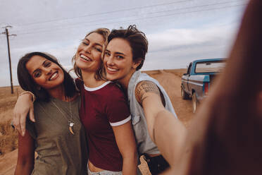 Gruppe von glücklichen jungen Frauen, die ein Selfie machen, während sie auf einem Roadtrip sind. Drei junge Freundinnen posieren für ein Selfie auf einer Landstraße mit einem Pickup-Truck im Hintergrund. - JLPSF24274